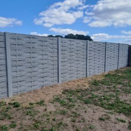 Wulkabet Producent Ogrodzeń Betonowych - Długi, szary, betonowy płot o wzorze plecionki, oddzielający teren. Widoczne słupki i panele. W tle błękitne niebo z chmurami i zieleń drzew.