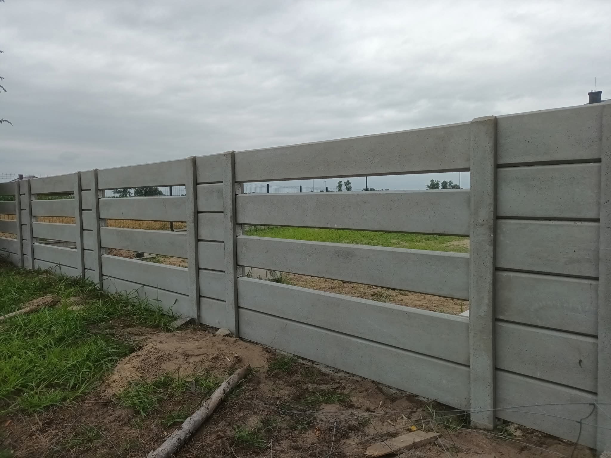 Szary, prefabrykowany płot betonowy z poziomymi szczelinami, widoczny z bliska na tle pochmurnego nieba i trawy. Elementy montażowe w betonie.