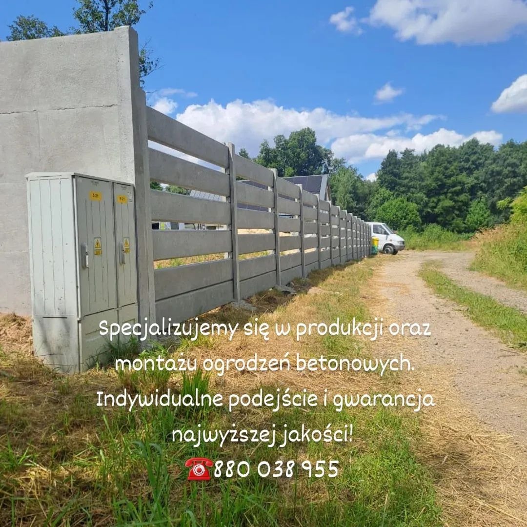 Długi, szary, betonowy płot panelowy z poziomymi szczelinami, biegnący wzdłuż polnej drogi, z zielenią i skrzynką elektryczną w tle, w słoneczny dzień.