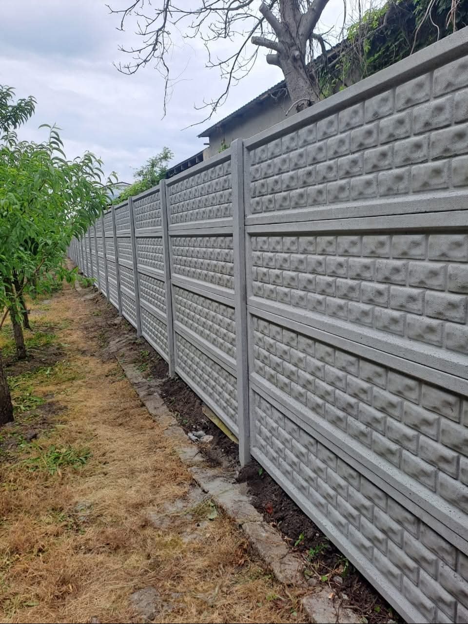 Długi, szary, betonowy płot z wzorem cegieł, ciągnący się wzdłuż trawnika z młodymi drzewkami. Widok z boku, pochmurne niebo w tle.