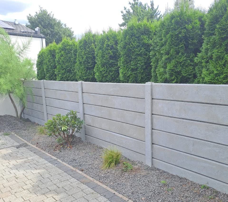 Szary, poziomy płot betonowy z roślinami i drzewami w tle, widziany z niskiej perspektywy. Płot oddziela posesję od zieleni, tworząc estetyczne ogrodzenie.