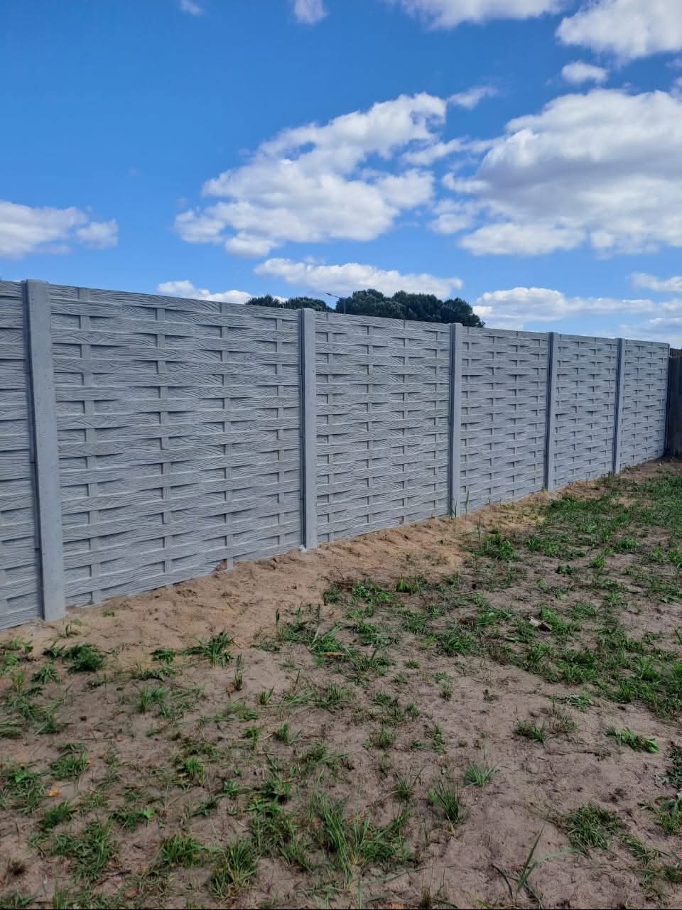 Długi, szary, betonowy płot o wzorze plecionki, oddzielający teren. Widoczne słupki i panele. W tle błękitne niebo z chmurami i zieleń drzew.