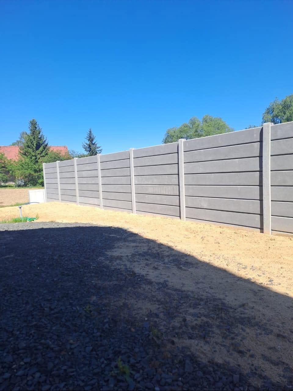 Nowoczesne ogrodzenie betonowe z poziomymi panelami, widoczne na tle błękitnego nieba i zieleni drzew. Częściowo zacienione podłoże z kamieniami i piaskiem.