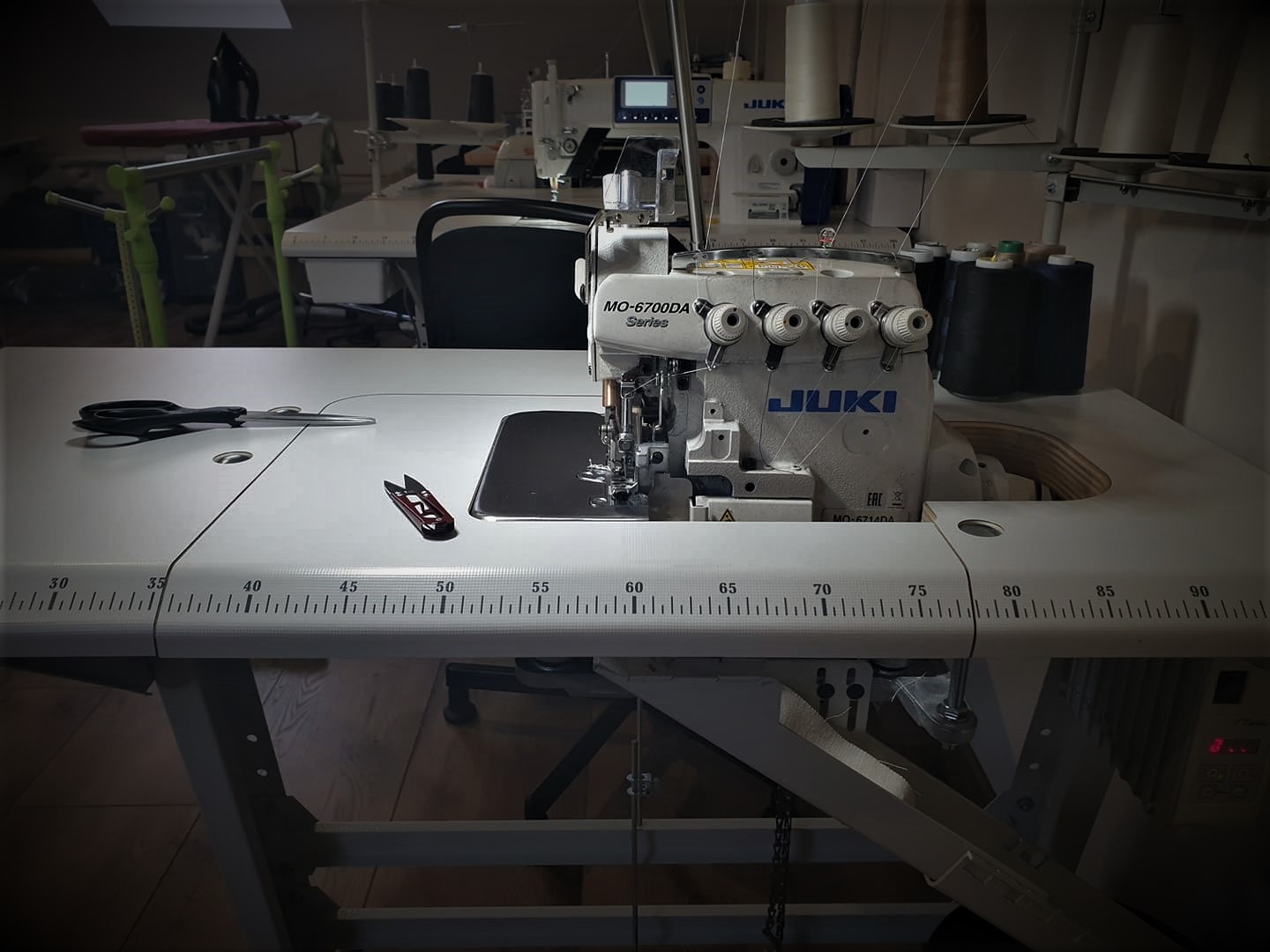 Overlock JUKI MO-6700DA na stole z miarką, nożyczki i obcinaczki leżą obok, w tle inne maszyny do szycia i nici.