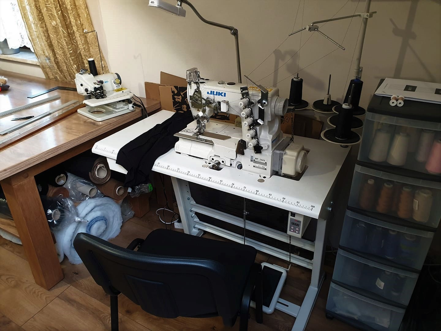 Przemysłowa maszyna do szycia JUKI MF-7523 na białym stole, obok druga mniejsza maszyna, w tle regał z nićmi i szpulami, widoczny fragment czarnego materiału.