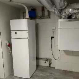 Dwa białe kotły grzewcze marki Vaillant zamontowane w pomieszczeniu wyłożonym białymi płytkami, widoczne rury i instalacje, na ścianie gniazdo elektryczne z czerwoną wtyczką.