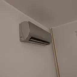 Zamontowany srebrny klimatyzator ścienny w jasnym pomieszczeniu z widocznymi rurami instalacyjnymi i oknem z niebieską taśmą malarską.