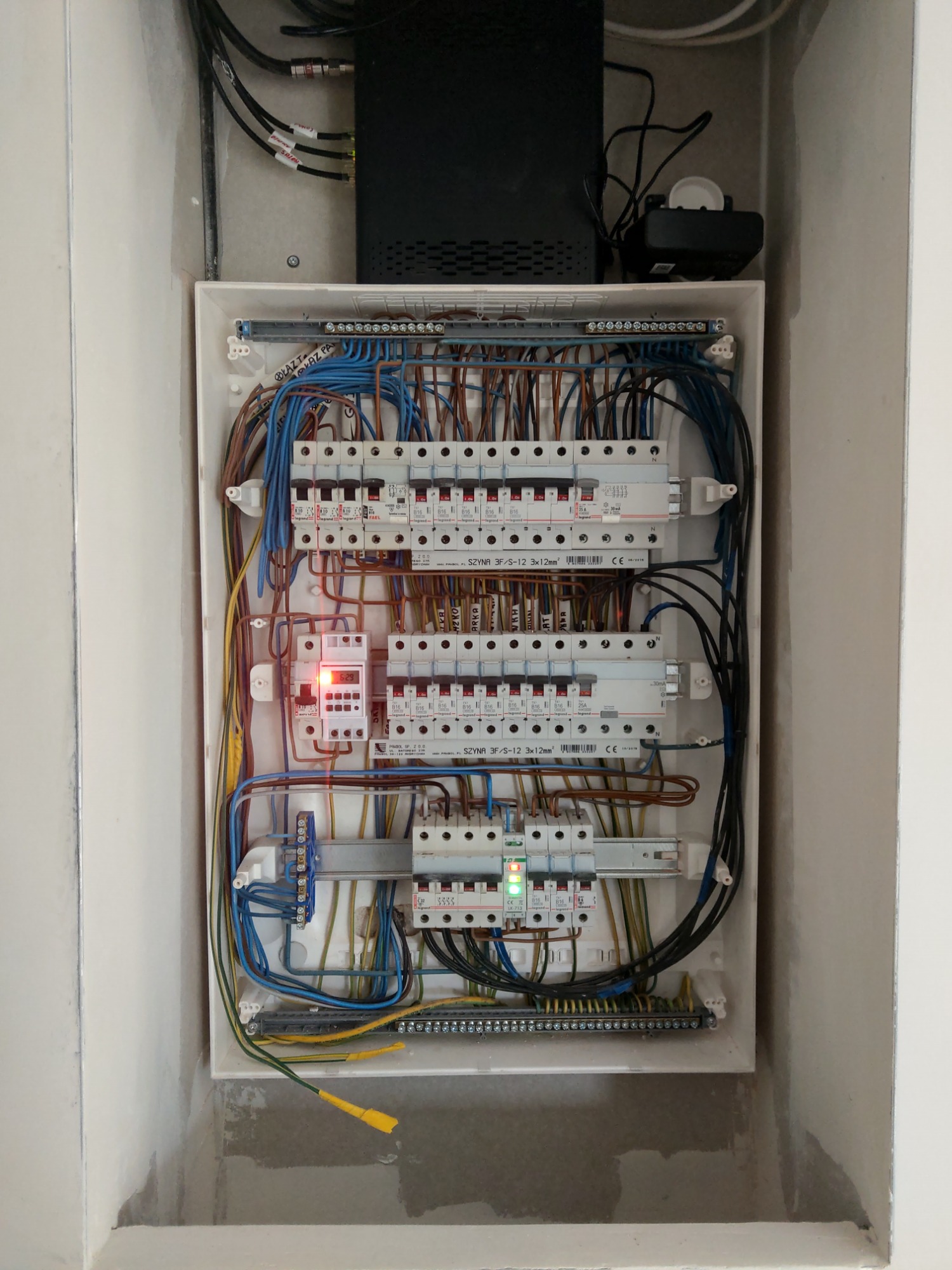 Otwarta skrzynka elektryczna z widocznymi bezpiecznikami, przewodami i elementami łączeniowymi, zamontowana we wnęce w ścianie, z dodatkowym urządzeniem elektronicznym na górze.