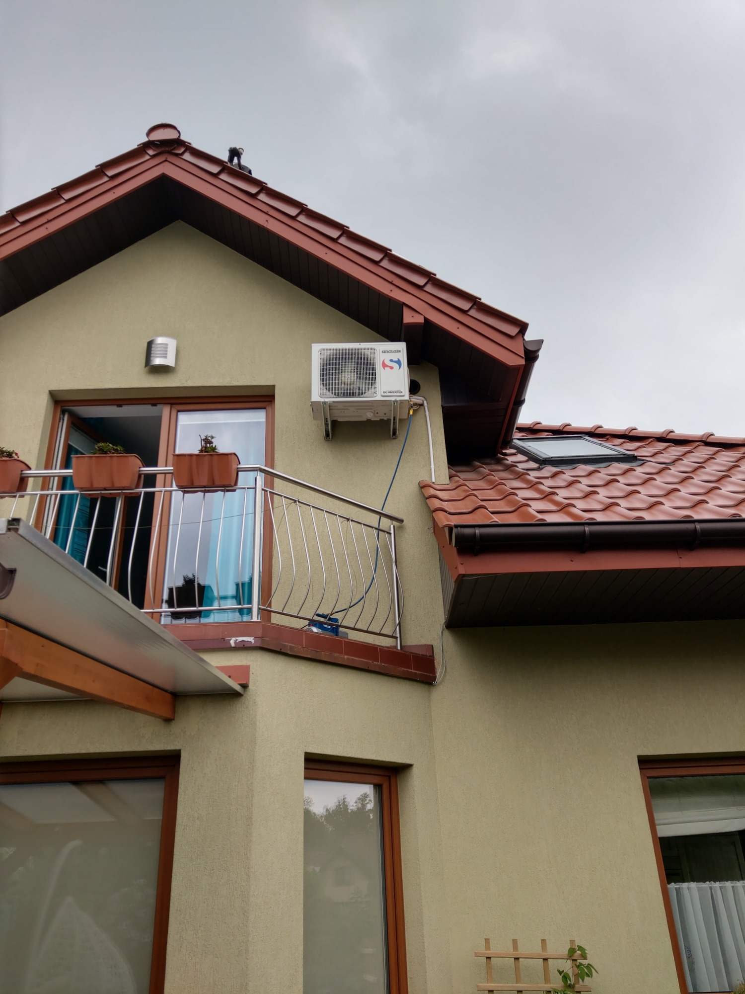 Biała jednostka zewnętrzna klimatyzatora zamontowana na ścianie domu z beżową elewacją, widoczny balkon z metalową balustradą i brązową obudową, czerwona dachówka i szare niebo w tle.