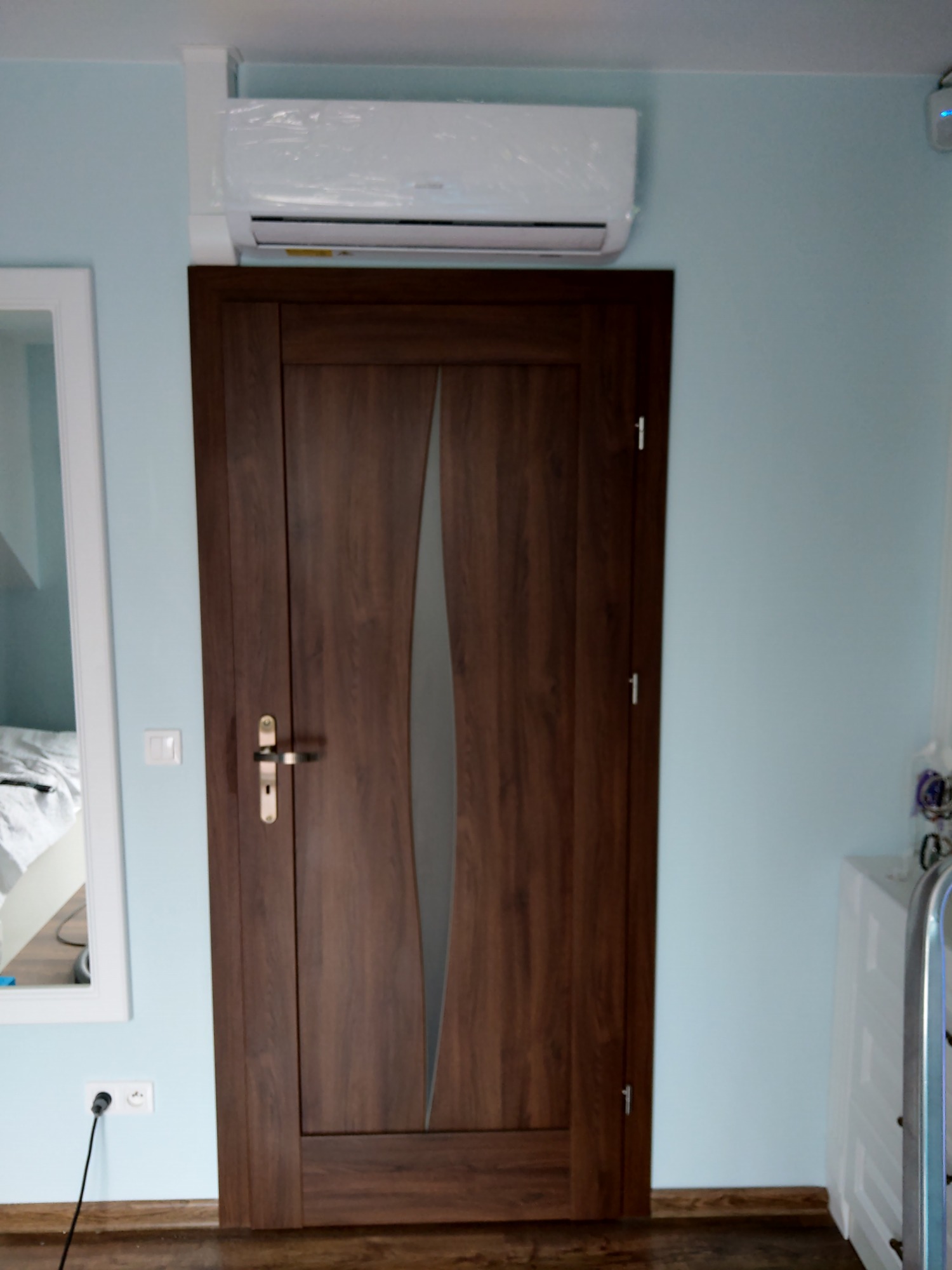 Zamontowany klimatyzator w folii ochronnej nad nowoczesnymi drzwiami wewnętrznymi w pokoju o jasnoniebieskich ścianach.