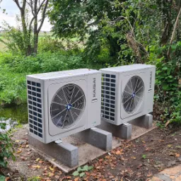 Dwie jednostki zewnętrzne pompy ciepła marki Kaisai, zamontowane na betonowych podstawach w otoczeniu zieleni i drzew, z widocznym fragmentem strumyka i pola w tle.