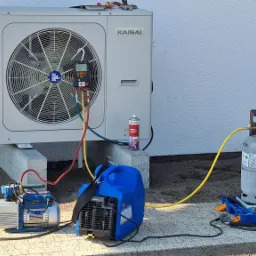 Biała jednostka zewnętrzna klimatyzacji Kaisai na betonowym fundamencie, podłączona do manometrów i butli z czynnikiem chłodniczym R-32, obok pompa próżniowa i niebieska sprężarka, podczas serwisu...