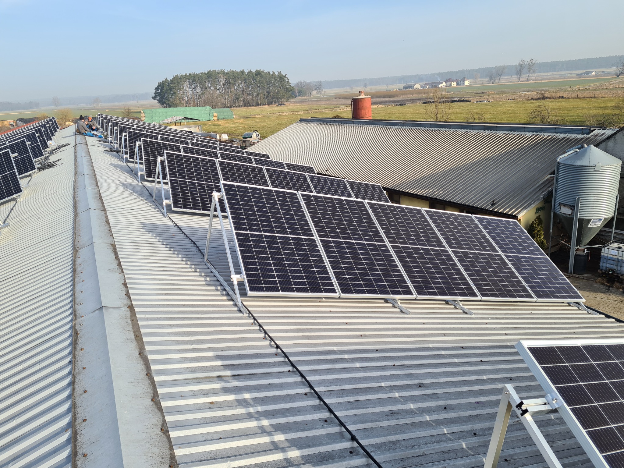 Rząd paneli słonecznych na blaszanym dachu budynku gospodarczego, w tle pola i las, dwóch monterów przy pracy.