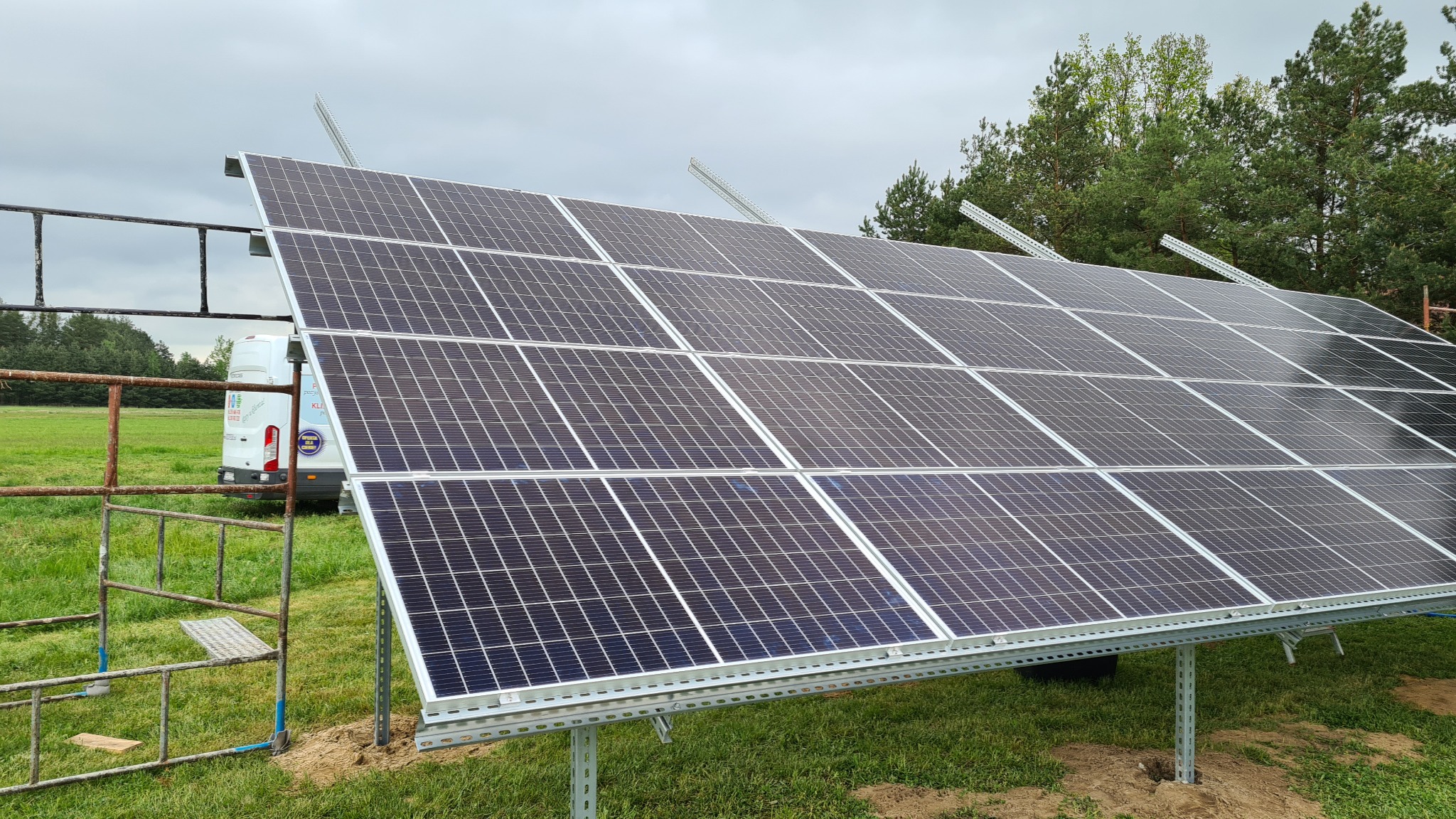 Panel słoneczny ustawiony na metalowej konstrukcji na trawiastym terenie, w tle widoczny samochód dostawczy i drzewa.
