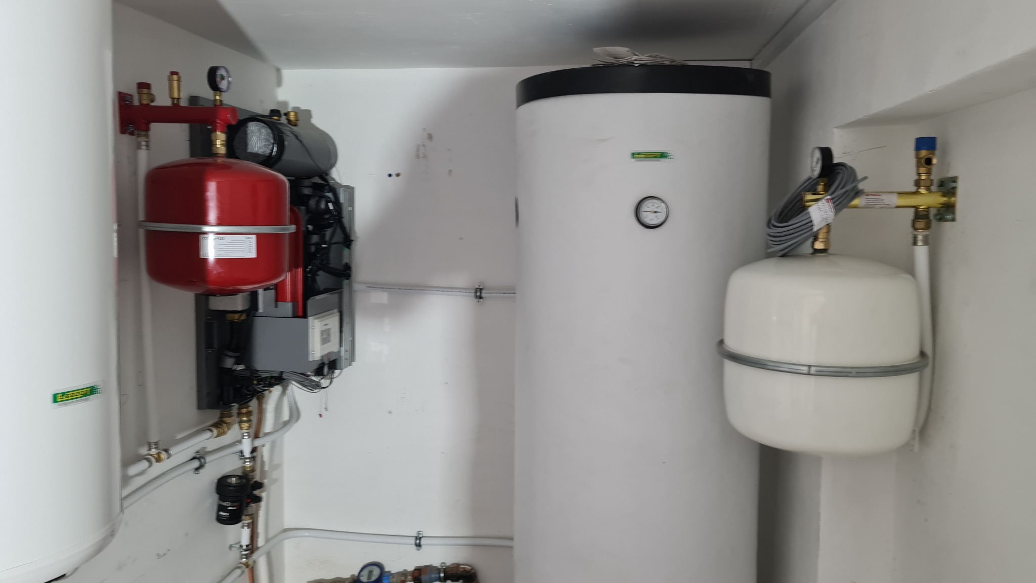Instalacja grzewcza w kotłowni: biały zbiornik buforowy z czarnym wiekiem, czerwony zbiornik wyrównawczy, szare rury i zawory w schludnym pomieszczeniu.