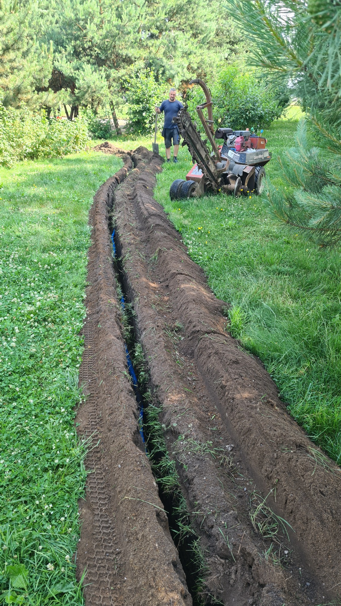 Wykopany rów w trawie z ułożoną niebieską rurą, wykonany przy użyciu glebogryzarki Ditch Witch, mężczyzna z łopatą w tle.