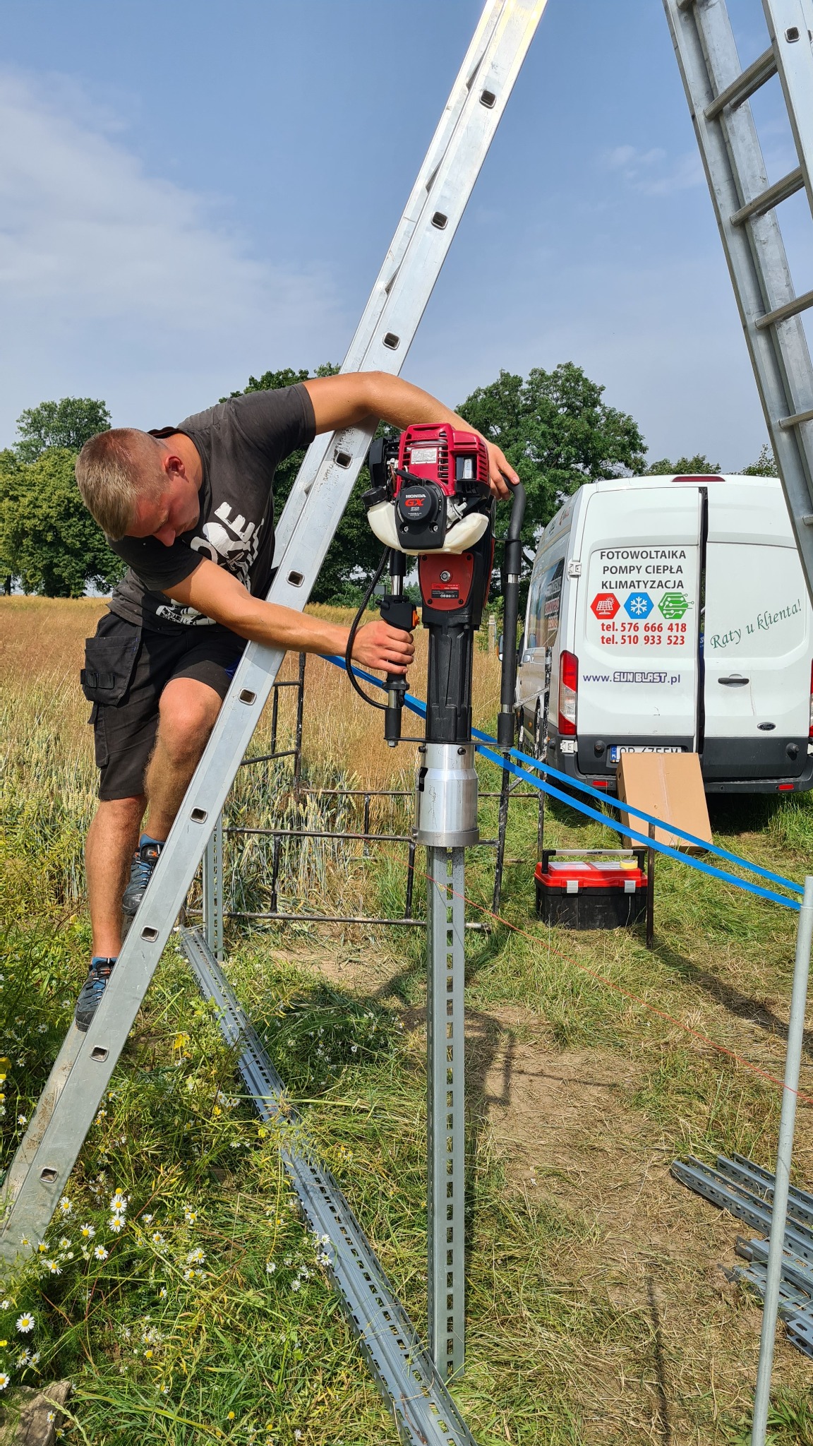 Monter na drabinie, wbija palik w ziemię za pomocą wbijaka spalinowego Honda GX35, w tle bus z reklamą firmy od fotowoltaiki, pomp ciepła i klimatyzacji