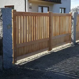 Nowoczesna, dwuskrzydłowa brama drewniana z jasnego drewna, zamontowana na granitowych słupkach, w Starym Sączu, z widocznymi cieniami na żwirowym podłożu.