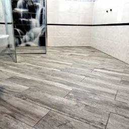 Wykończona łazienka z płytkami imitującymi drewno na podłodze ułożonymi pod kątem, białe płytki na ścianach z czarnym pasem dekoracyjnym, kabina prysznicowa z grafiką wodospadu.