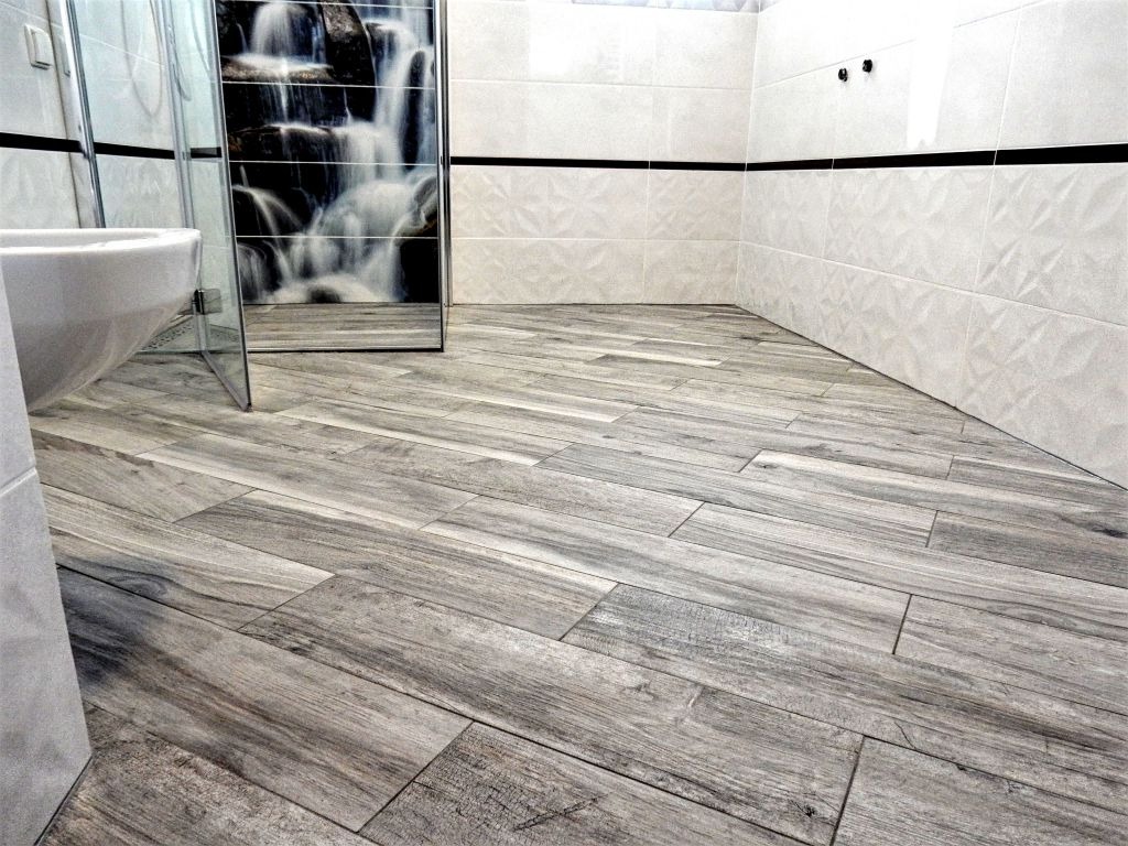 Wykończona łazienka z płytkami imitującymi drewno na podłodze ułożonymi pod kątem, białe płytki na ścianach z czarnym pasem dekoracyjnym, kabina prysznicowa z grafiką wodospadu.