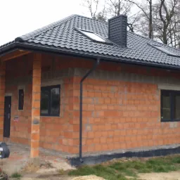 Dom w trakcie budowy z cegły, z czarną dachówką, oknami i rynnami. Na pierwszym planie betoniarka. Widoczne drzewa w tle.