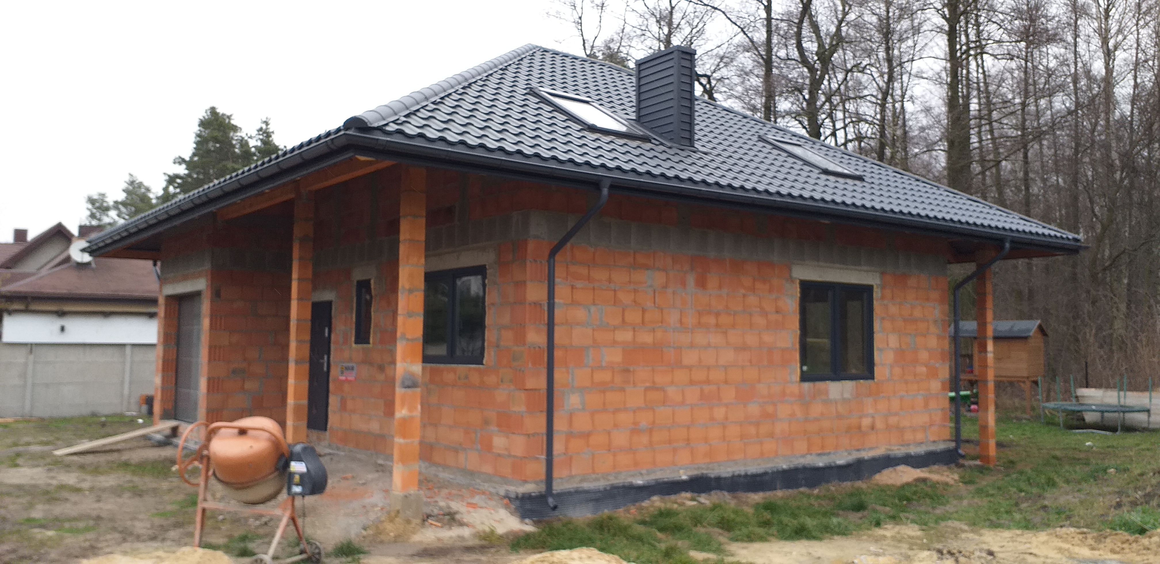 Dom w trakcie budowy z cegły, z czarną dachówką, oknami i rynnami. Na pierwszym planie betoniarka. Widoczne drzewa w tle.