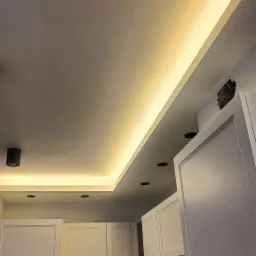 Sufit z podwieszanym oświetleniem LED wzdłuż krawędzi, widoczne również czarne reflektory punktowe i białe szafki kuchenne z mozaiką na ścianie.