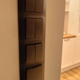 Panel z czterema włącznikami światła w kolorze ciemnego brązu, zamontowany na jasnej ścianie, widoczny fragment białej zabudowy kuchennej w tle.