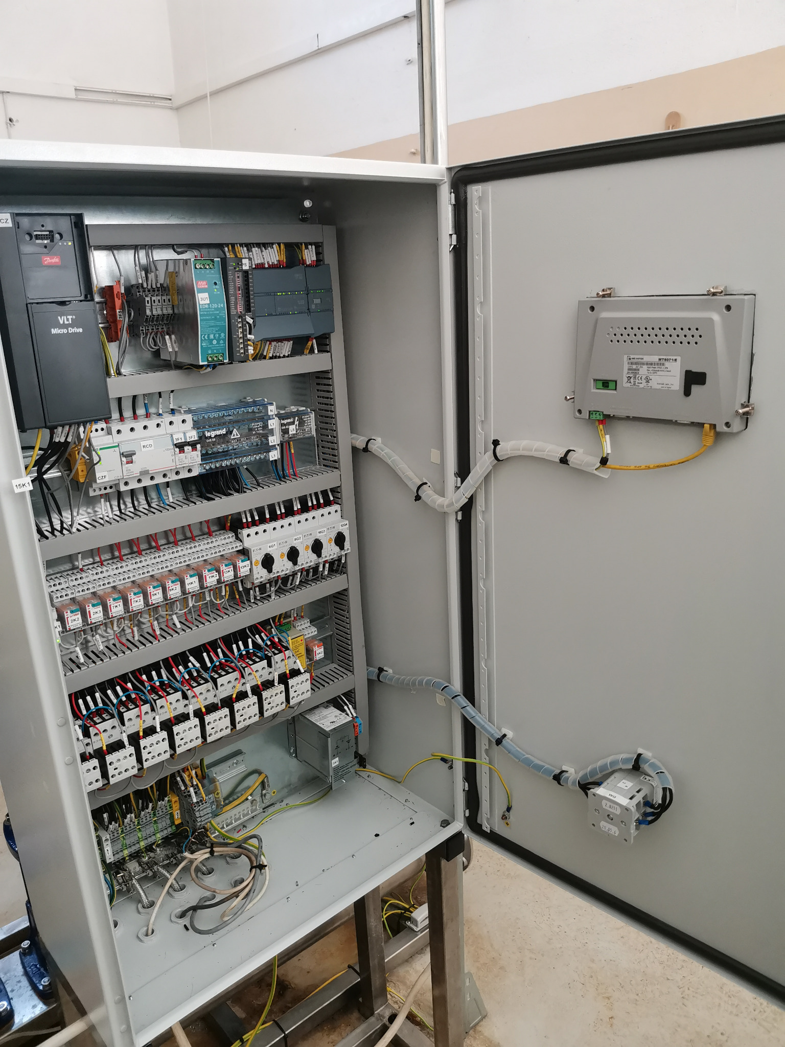 Otwarta szafa sterownicza z modułami elektronicznymi, bezpiecznikami i okablowaniem, widoczne elementy sterujące i zasilające, kable prowadzone w peszlach. Na drzwiach szafy zamontowany panel...