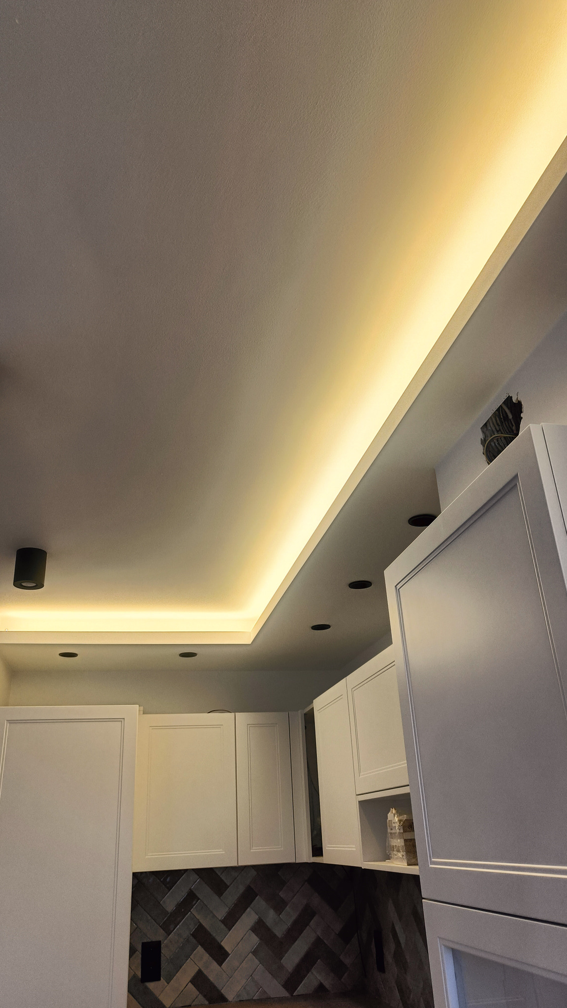 Sufit z podwieszanym oświetleniem LED wzdłuż krawędzi, widoczne również czarne reflektory punktowe i białe szafki kuchenne z mozaiką na ścianie.