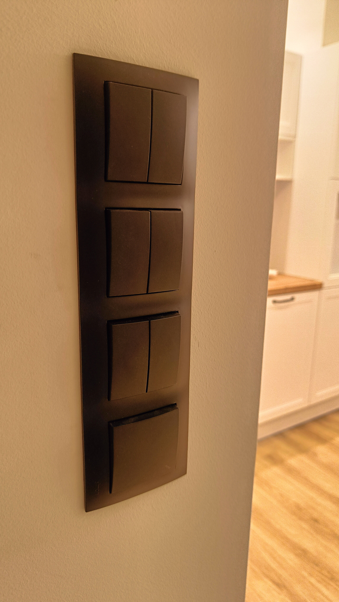 Panel z czterema włącznikami światła w kolorze ciemnego brązu, zamontowany na jasnej ścianie, widoczny fragment białej zabudowy kuchennej w tle.