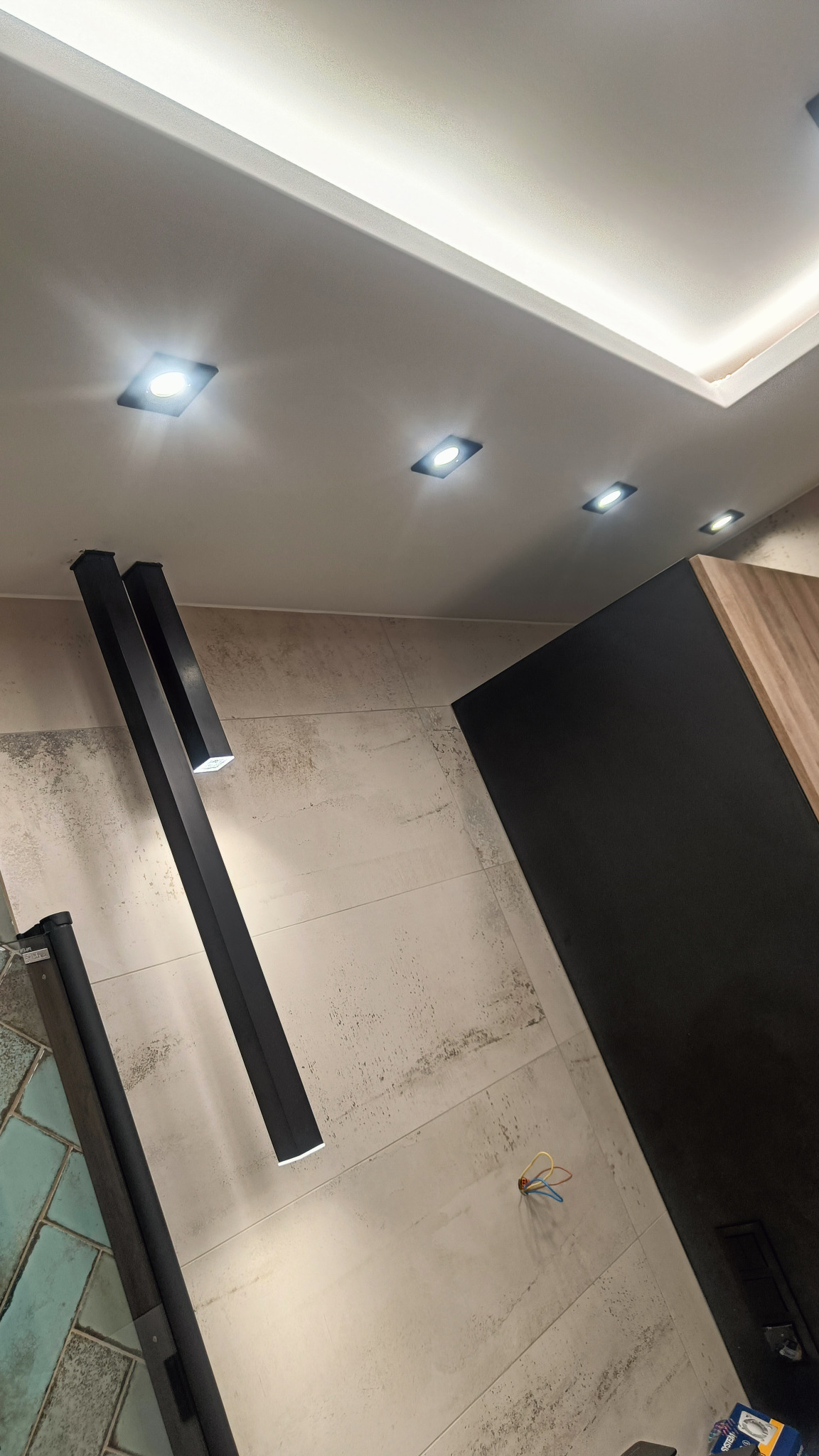 Nowoczesne oświetlenie sufitowe z czarnymi, podłużnymi lampami wiszącymi i wbudowanymi punktami świetlnymi, widoczne przewody elektryczne na ścianie.