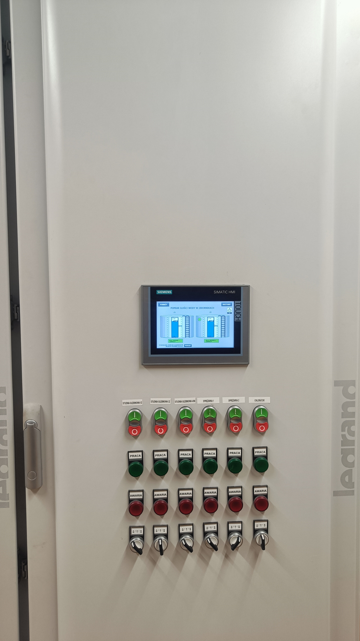 Panel sterowania Siemens Simatic HMI z wyświetlaczem graficznym i przyciskami sygnalizującymi pracę, awarię oraz przełącznikami, zamontowany w białej szafie sterowniczej Legrand.