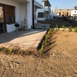 Nowo założony ogród z wyrównanym terenem, świeżo posadzonymi tujami wzdłuż ogrodzenia i wybrukowanym tarasem z grillem i huśtawką w tle.