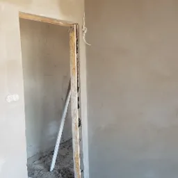 Świeżo otynkowane ściany w surowym wnętrzu, widoczne drewniane ościeżnice drzwiowe i wystające przewody elektryczne na podłodze.