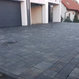 Nowoczesny podjazd z szarej kostki brukowej przed domem z dwoma garażami i reklamą dewelopera w tle.