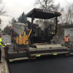 Rozściełacz asfaltu Volvo układa świeżą nawierzchnię drogową, z tyłu wywrotka z ładunkiem, pracownicy w odblaskowych ubraniach kontrolują proces, pochmurny dzień.