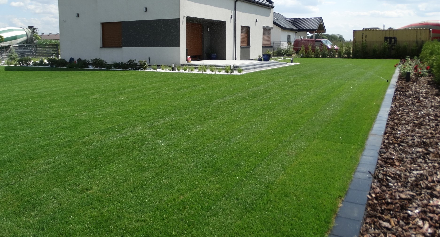Rozległy, intensywnie zielony trawnik przed nowoczesnym domem z tarasem wyłożonym jasnymi płytami, obrzeża z białego kamienia, w tle betoniarka i kontener.