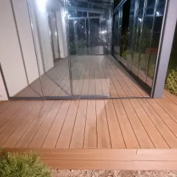 Zadaszony taras z desek kompozytowych, oświetlony nocą, z widocznymi elementami szklanej zabudowy i zieleni wokół.