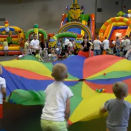 Dzieci bawią się kolorową chustą animacyjną na tle dmuchanych zamków i animatorów w strojach postaci z bajek podczas imprezy w hali.