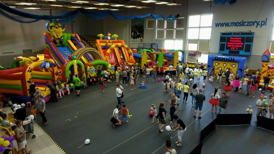 Hala sportowa wypełniona dmuchanymi zamkami i zjeżdżalniami dla dzieci, z motywami Minionków i palm, otoczona bawiącymi się dziećmi i dorosłymi.
