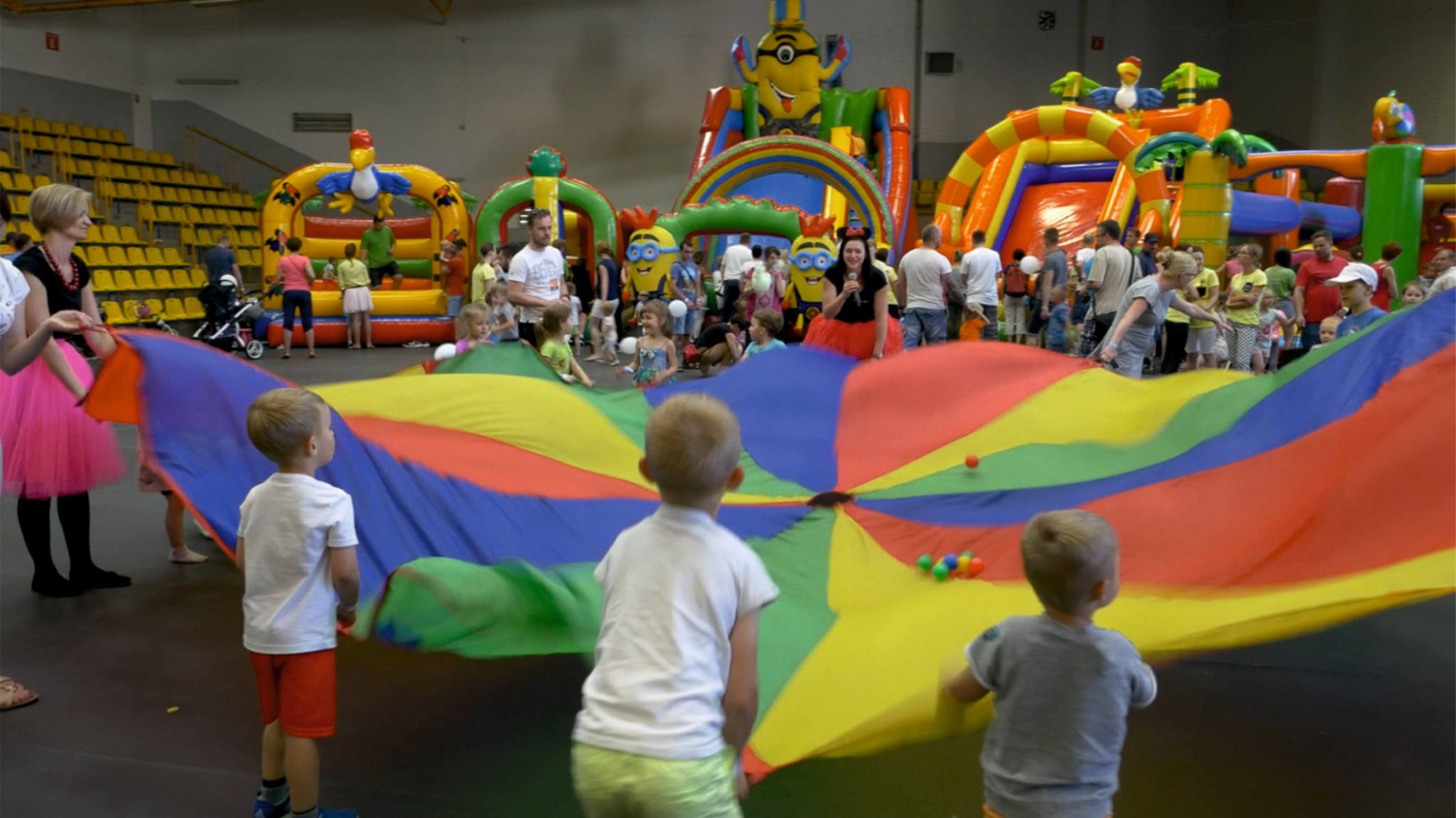 Dzieci bawią się kolorową chustą animacyjną na tle dmuchanych zamków i animatorów w strojach postaci z bajek podczas imprezy w hali.