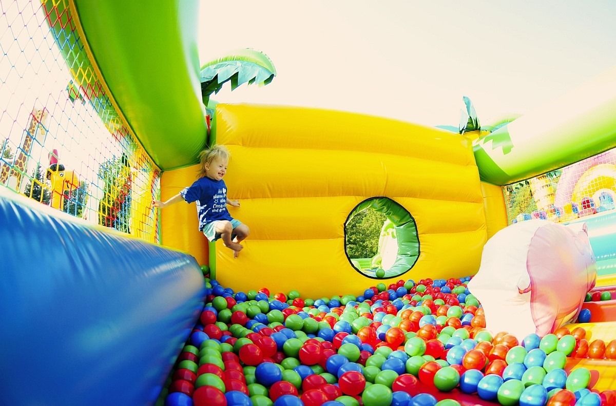 Uśmiechnięty chłopiec w niebieskiej koszulce skacze w kolorowym dmuchanym zamku z basenem wypełnionym plastikowymi kulkami.