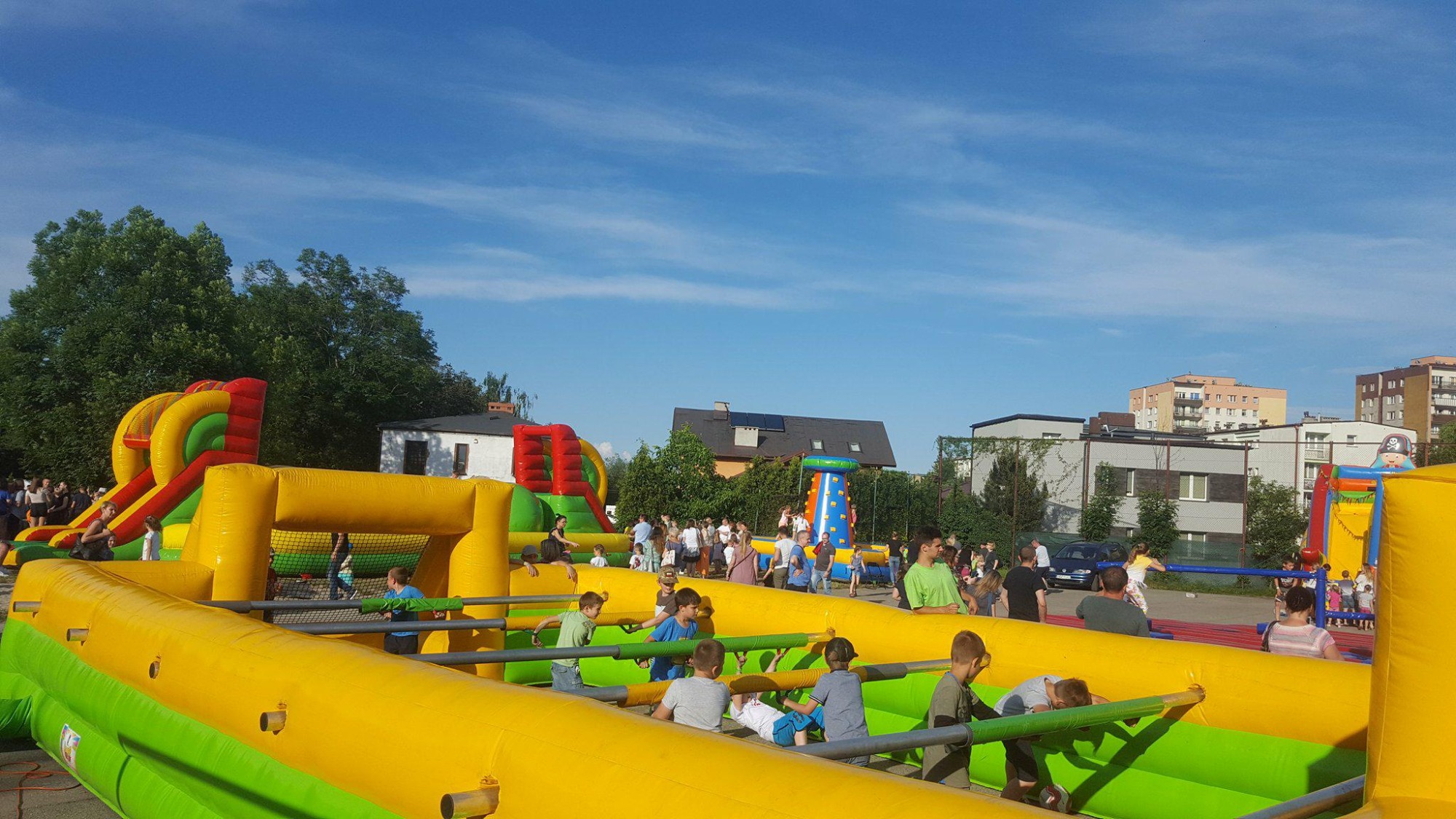 Dzieci bawiące się na dmuchanym boisku do piłkarzyków w plenerze, w tle dmuchana zjeżdżalnia i ścianka wspinaczkowa, widoczni dorośli i budynki mieszkalne.
