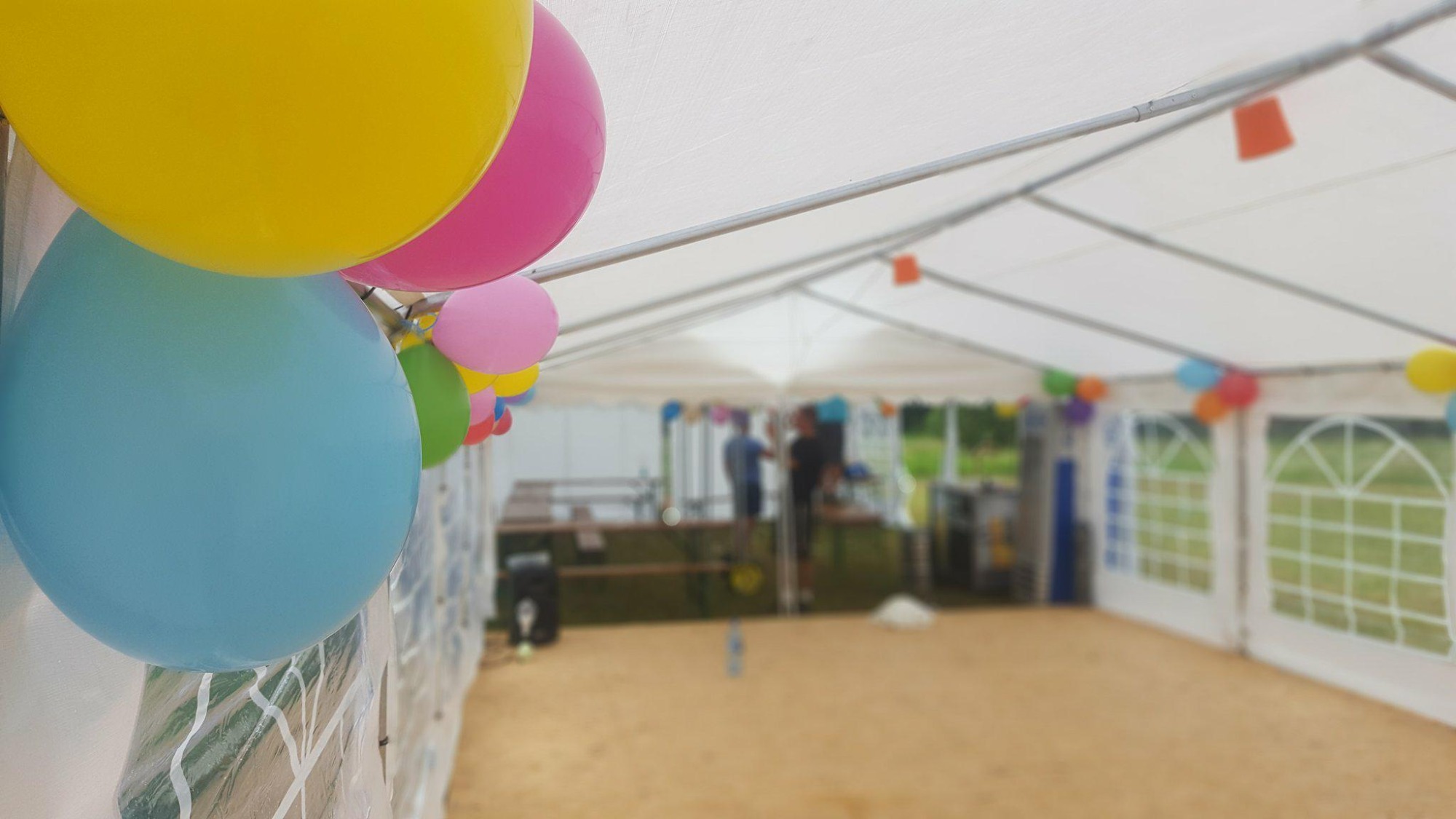 Wnętrze białego namiotu imprezowego udekorowanego kolorowymi balonami, widoczne ławostoły i osoby w tle.