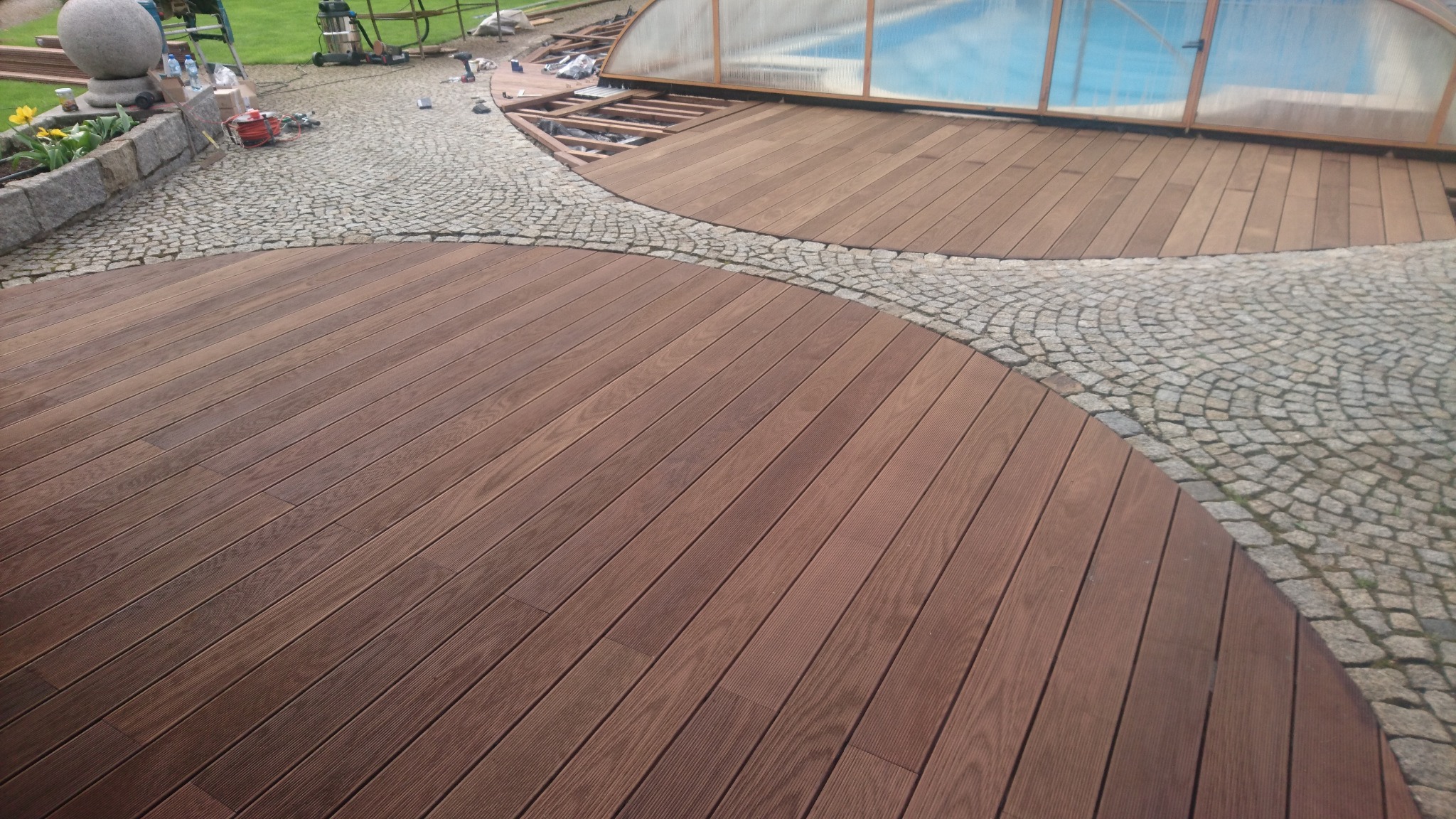 Brązowa deska tarasowa ułożona w łuk, oddzielona od pokrytej kostką brukową ścieżki, z widocznym fragmentem basenu ogrodowego w tle.