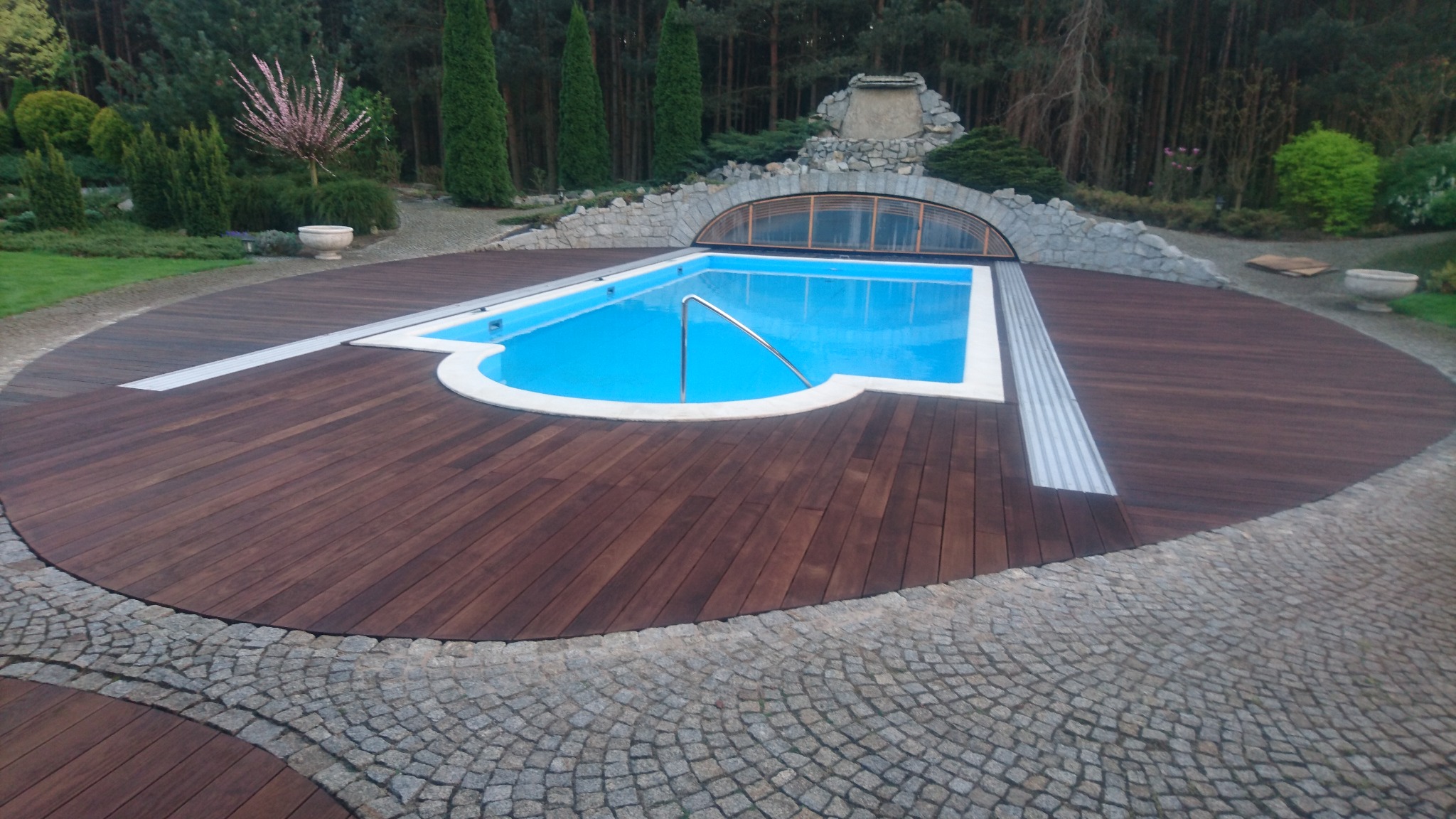 Okrągły taras z ciemnego drewna otaczający basen z błękitną wodą, z automatycznie otwieraną pokrywą i kamienną obudową w tle, otoczony zielenią.