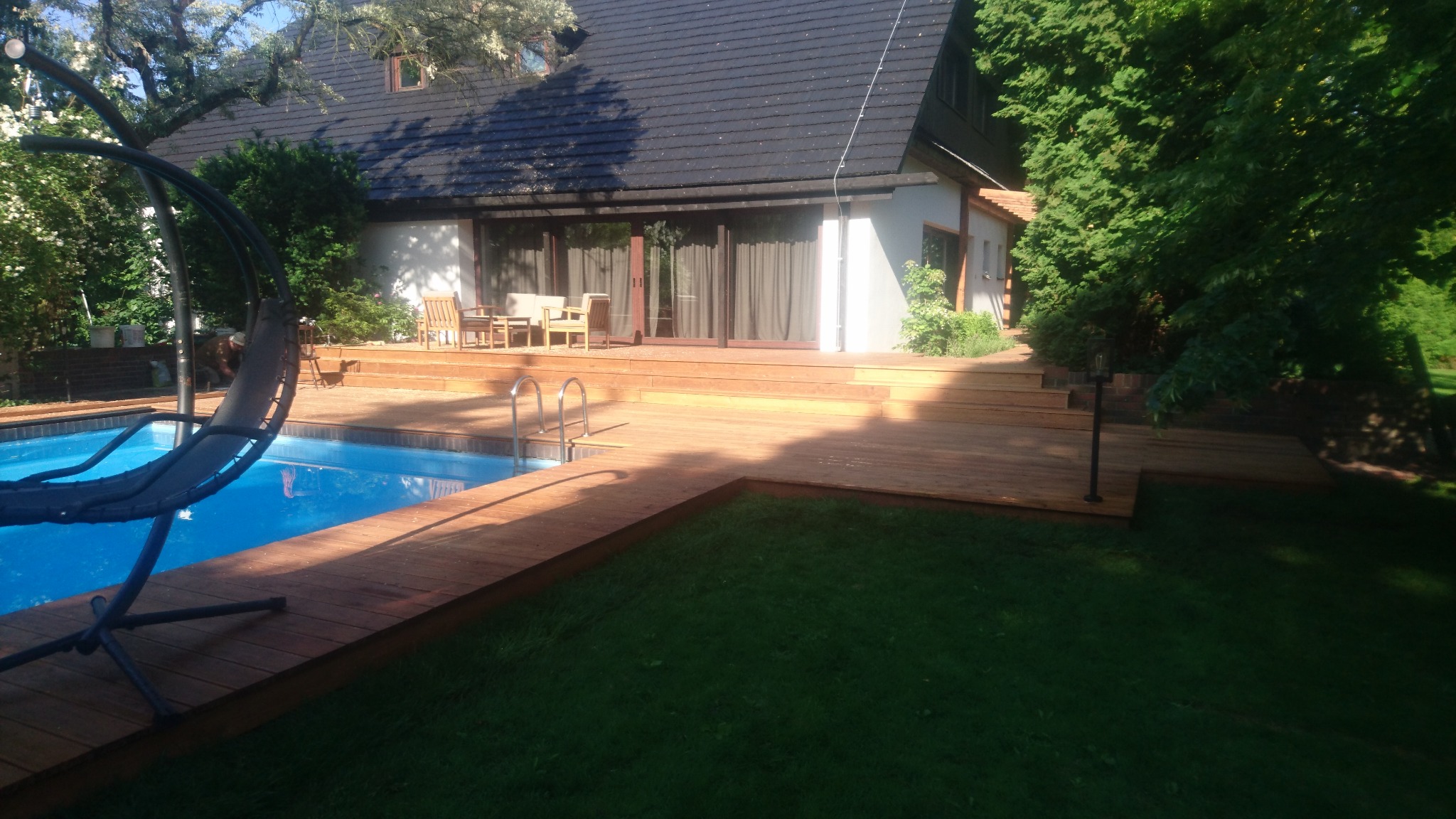 Przydomowy basen otoczony drewnianym tarasem z zejściem schodami, w tle dom z dużym oknem tarasowym i meblami ogrodowymi.