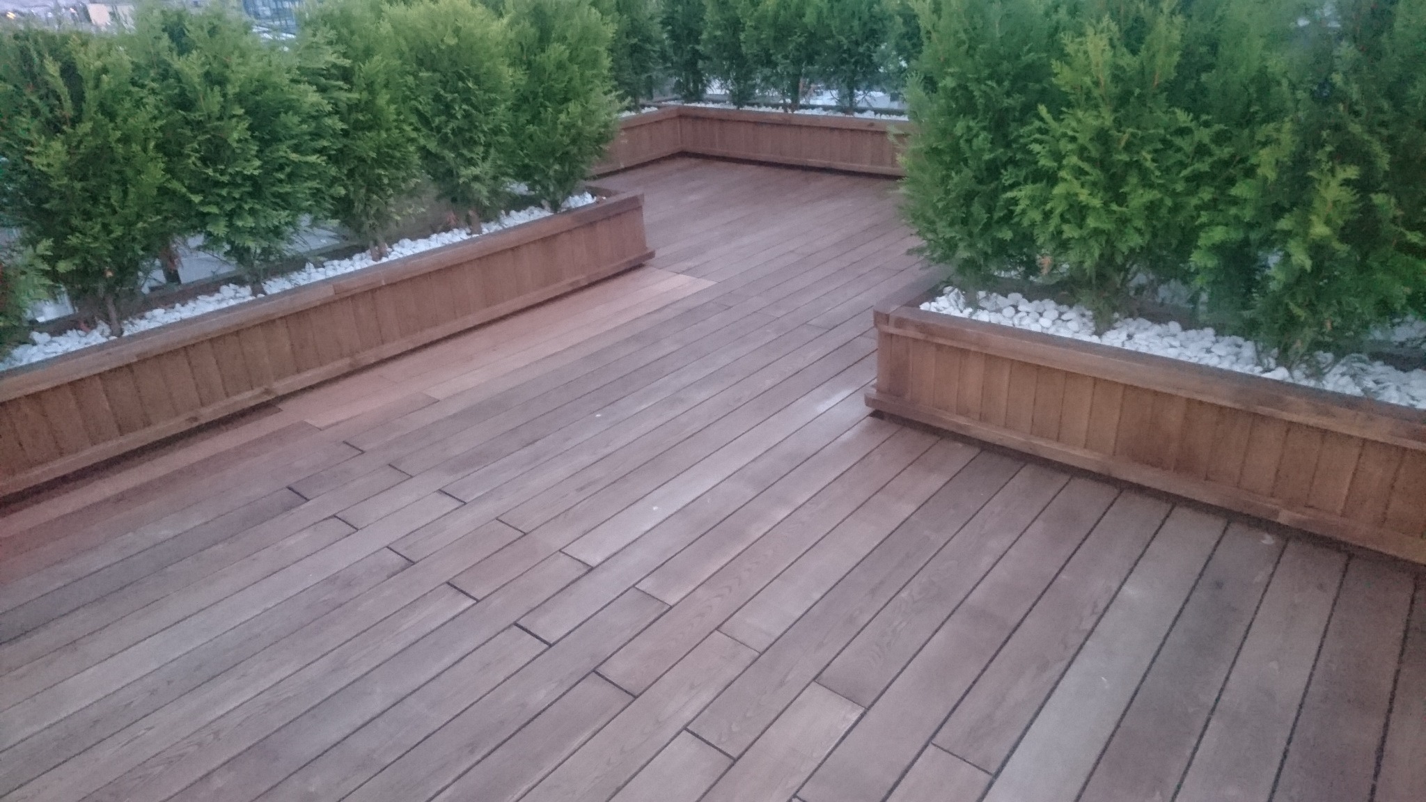 Drewniany taras z prostokątnymi deskami ułożonymi równolegle, obrzeża z drewnianymi donicami wypełnionymi białymi kamieniami i zielonymi krzewami.