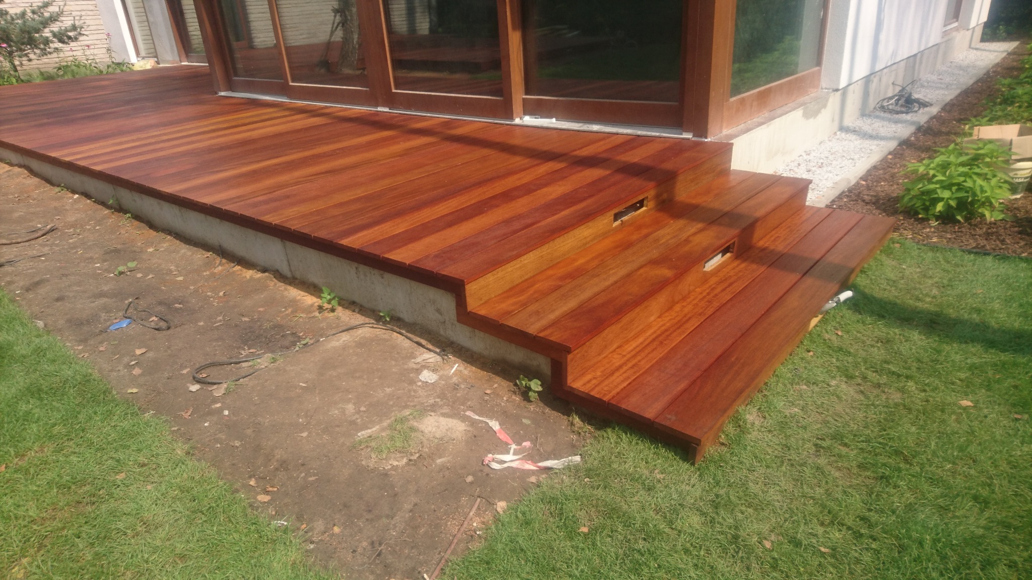 Nowo wybudowany taras z drewna egzotycznego z dwustopniowymi schodami, przylegający do budynku z dużymi przesuwnymi drzwiami, widoczne niedoskonałości terenu po budowie.